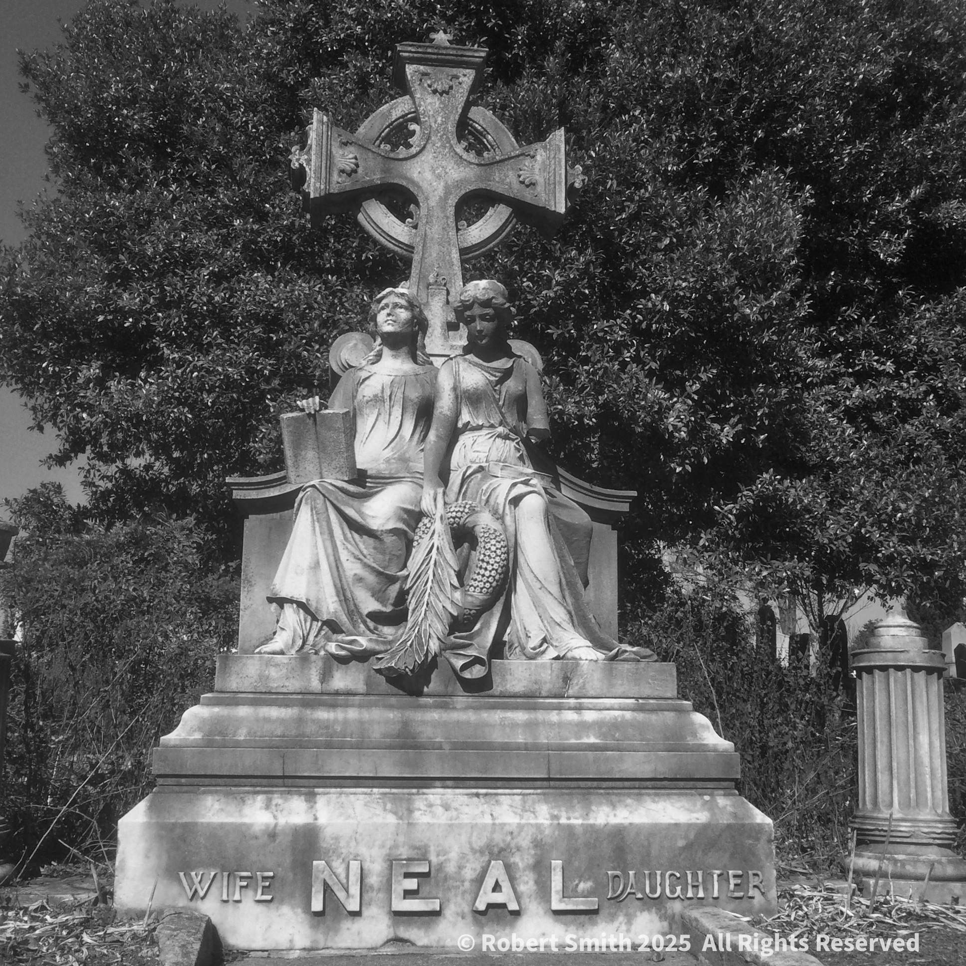 Black-and-white photograph of the NEAL family monument, with two seated stone figures beneath a large cross and “Wife” and “Daughter” carved at its base.