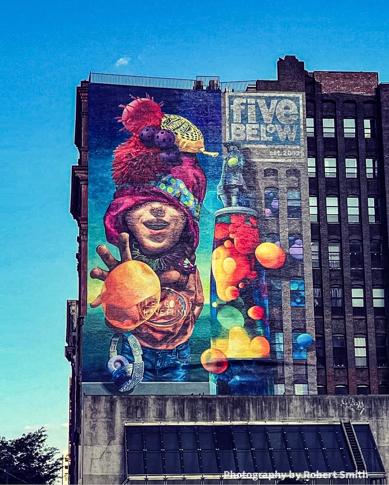 Frontal view of the Five Below mural with a smiling child in a pink hat, an outstretched hand, colorful floating shapes, and a tall lava lamp.