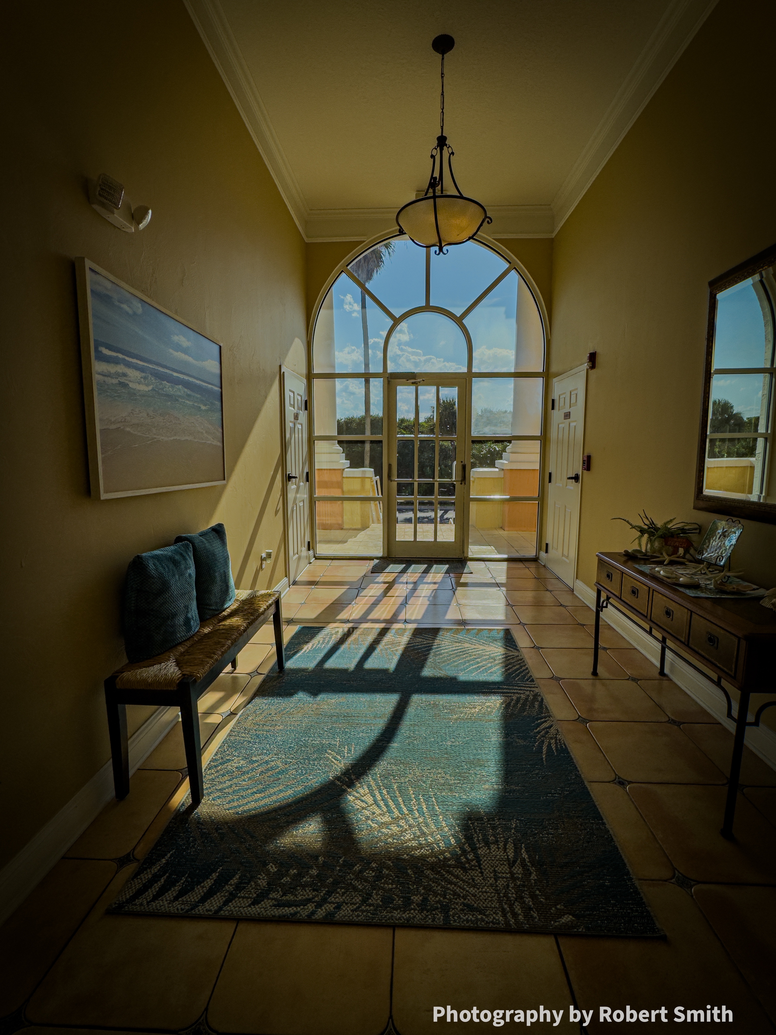 Tall arched glass doorway at the end of a warm-toned entry hall, casting long window shadows across a blue-green rug while bright sky shows outside.