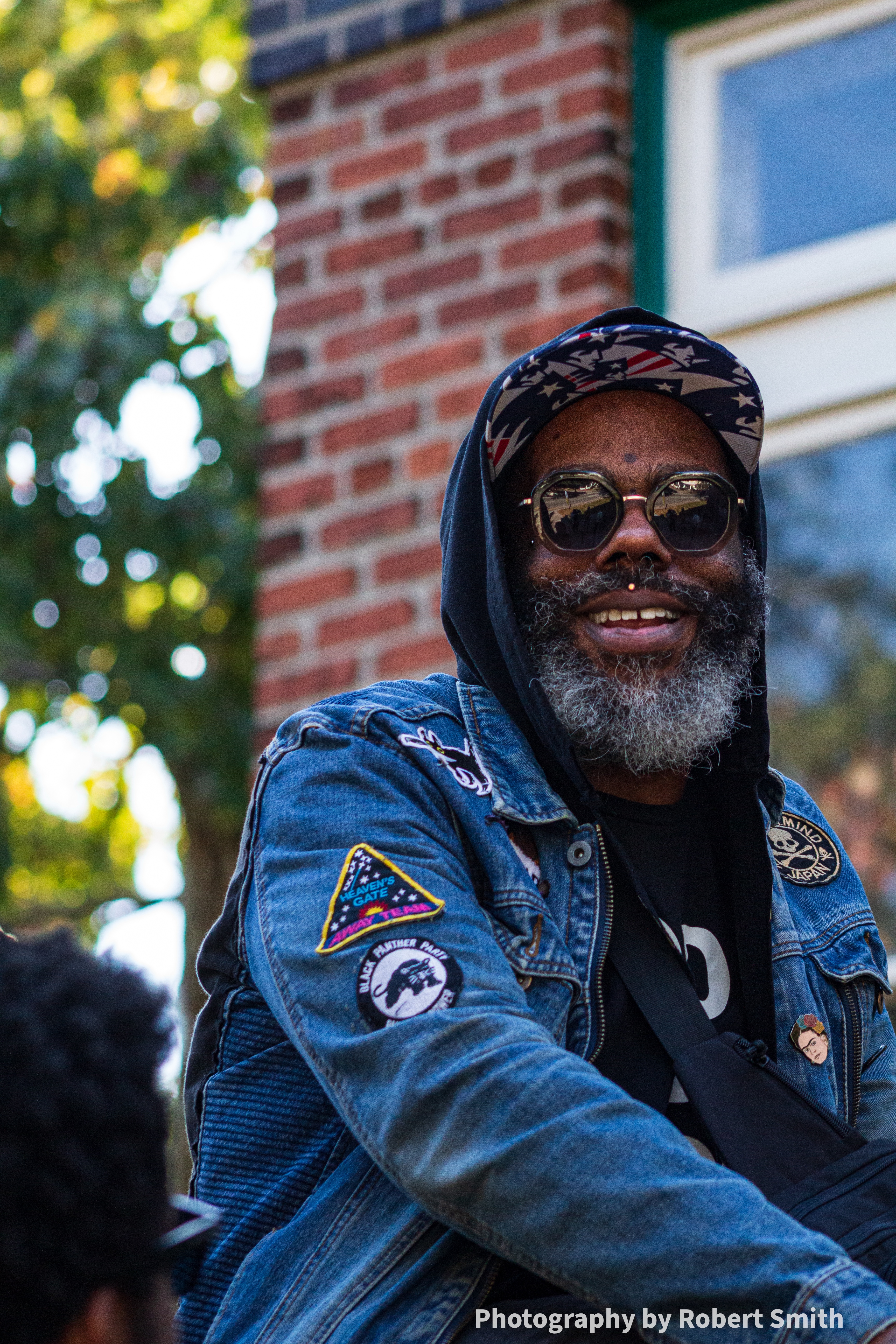 Smiling Black man seated outdoors in sunglasses, hood, patterned cap, and denim jacket near a historic brick building.