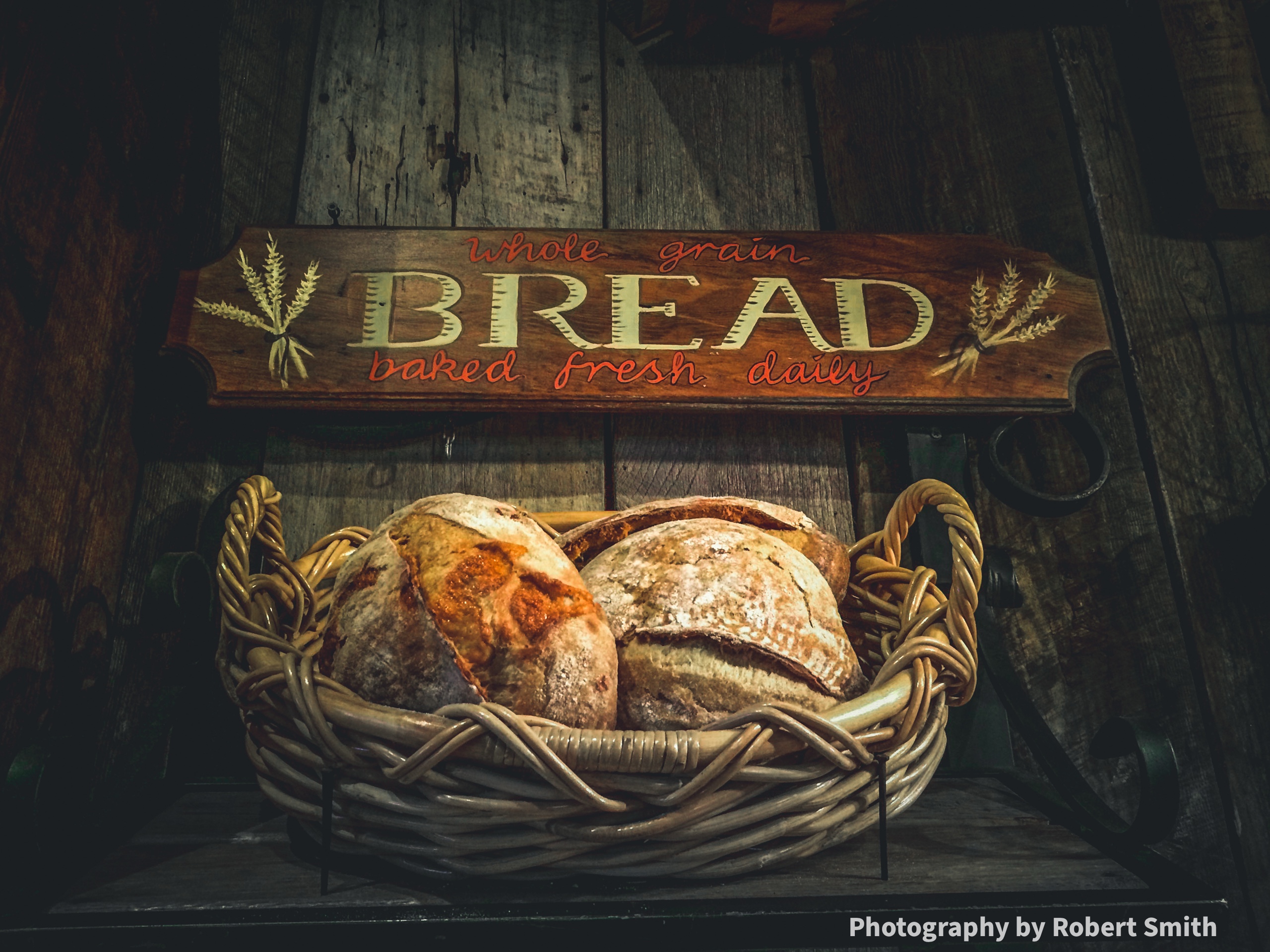 A wicker basket filled with rustic loaves beneath a wooden sign reading “Whole Grain Bread, Baked Fresh Daily” against a dark, weathered wood wall.