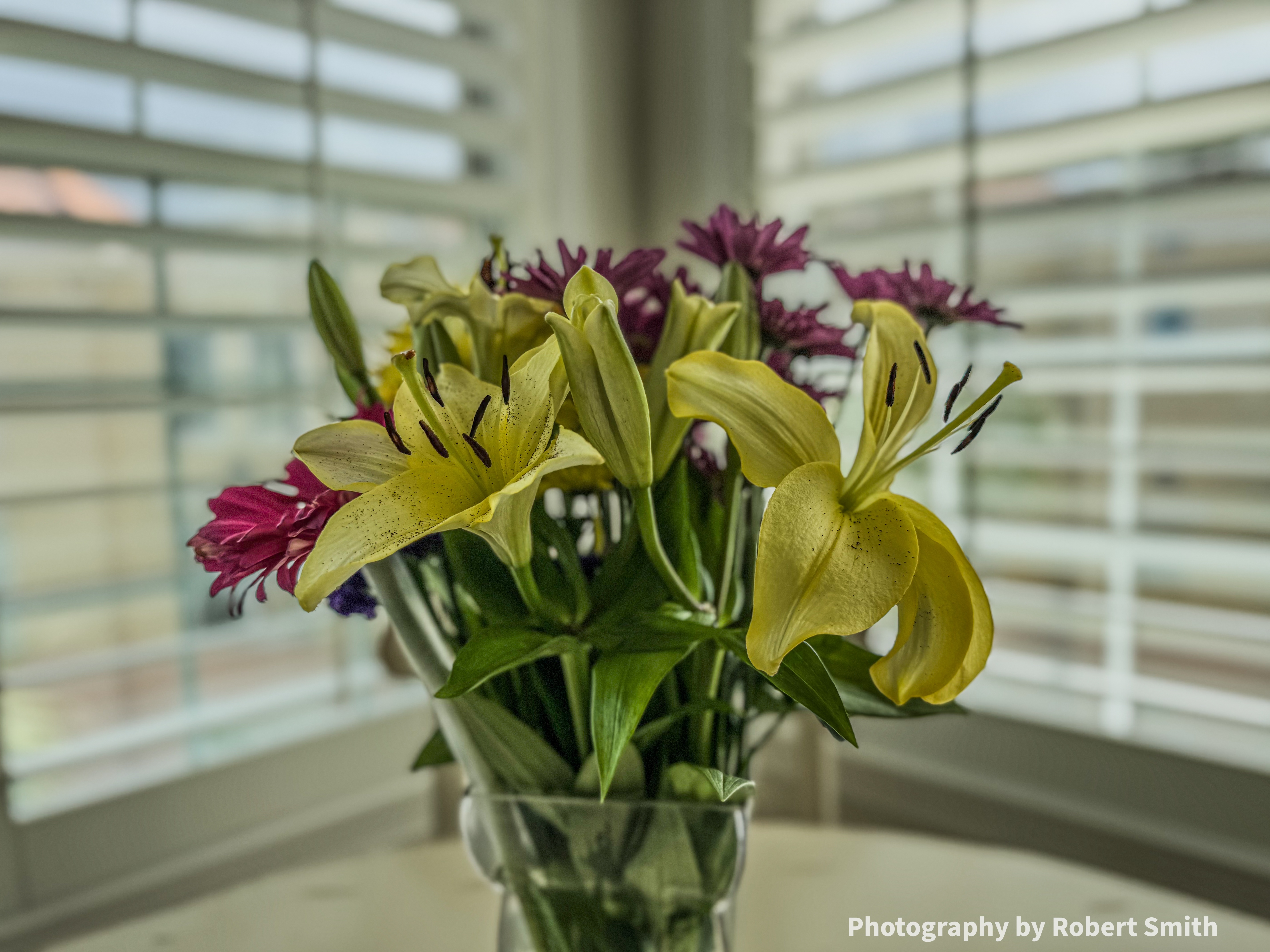 Two open yellow lilies with speckled petals and dark stamens fill the foreground of a mixed bouquet in a clear glass vase beside white shutters.