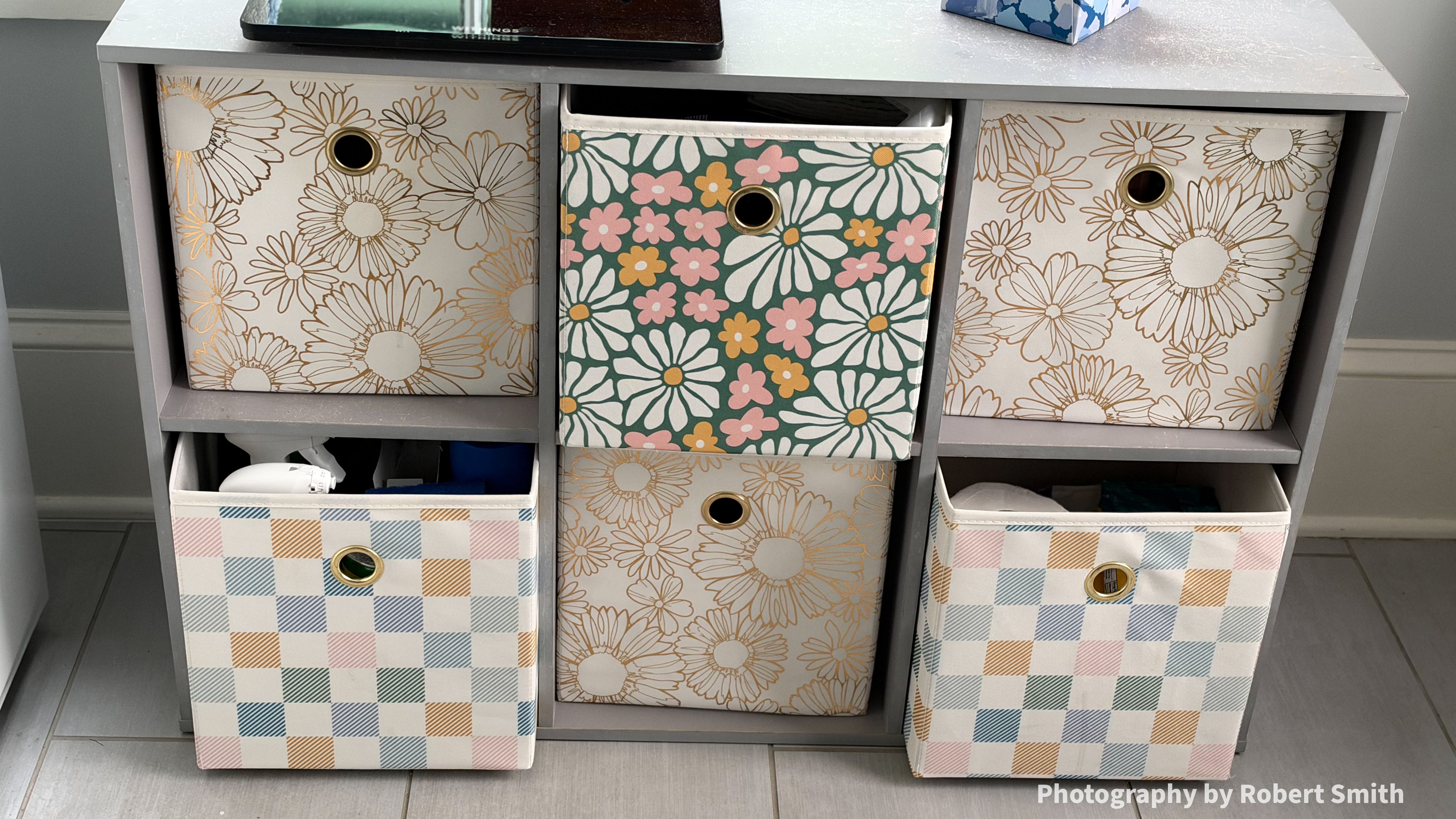 Close view of six patterned storage cubes in gray shelving, with the green floral cube and two pastel checked cubes pulled forward, revealing stored bathroom items.