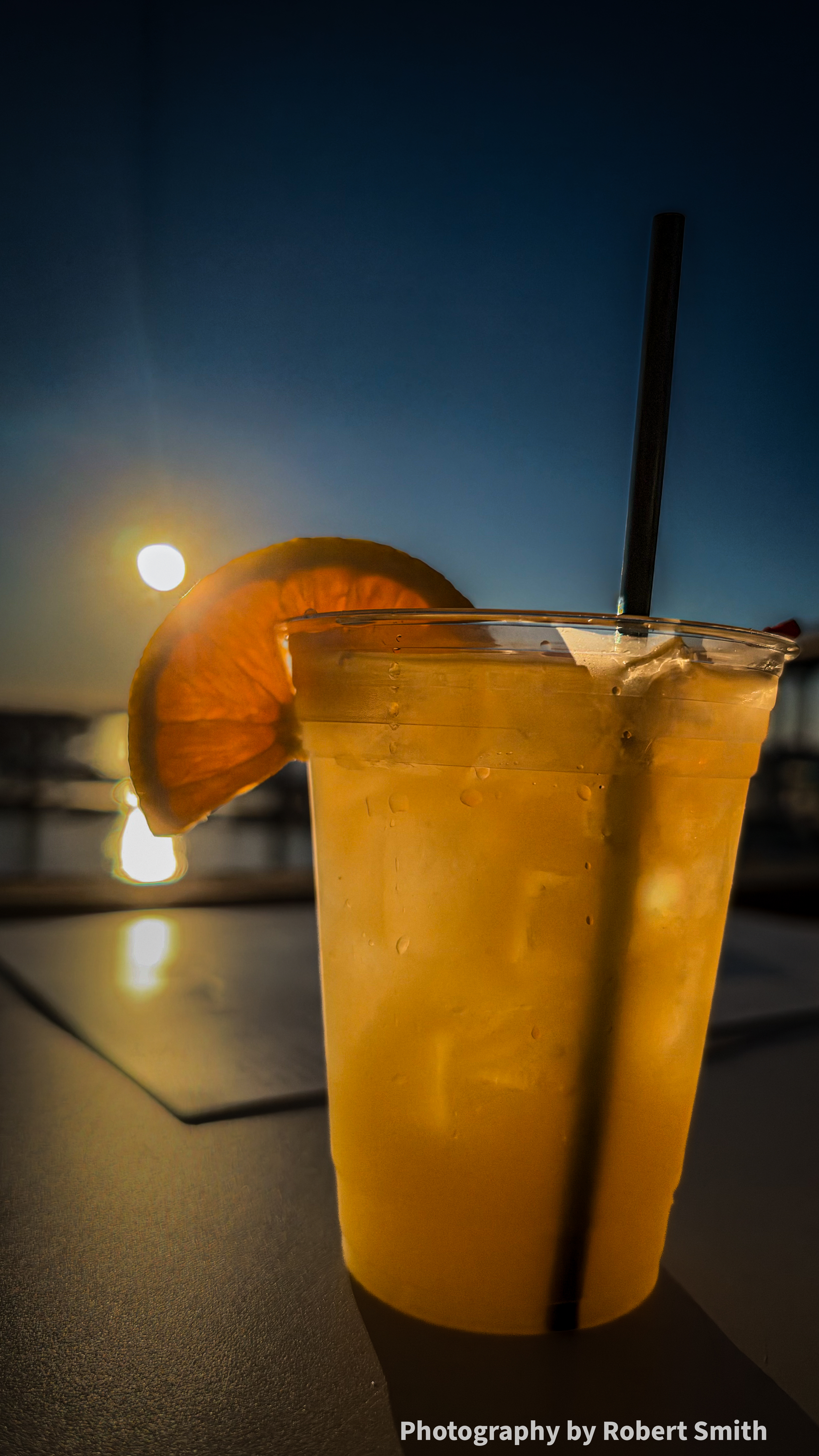 Close view of a golden whisky sour with condensation on the cup and a backlit orange slice, with the low sun above the garnish and blurred river reflections behind it.