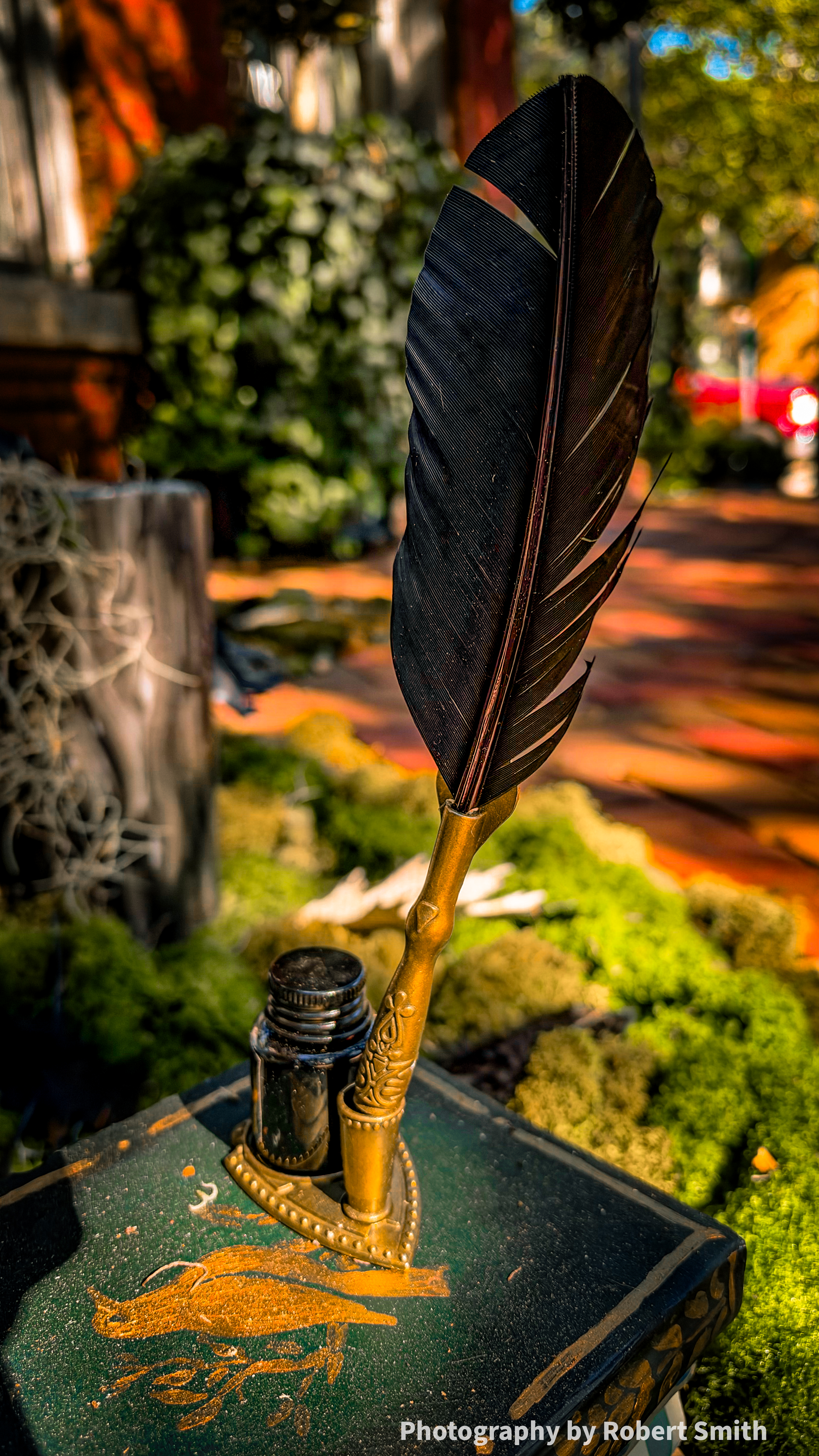 Close view of a black feather quill, ornate gold-colored holder, and dark inkwell atop a green decorative book with a gold bird design.