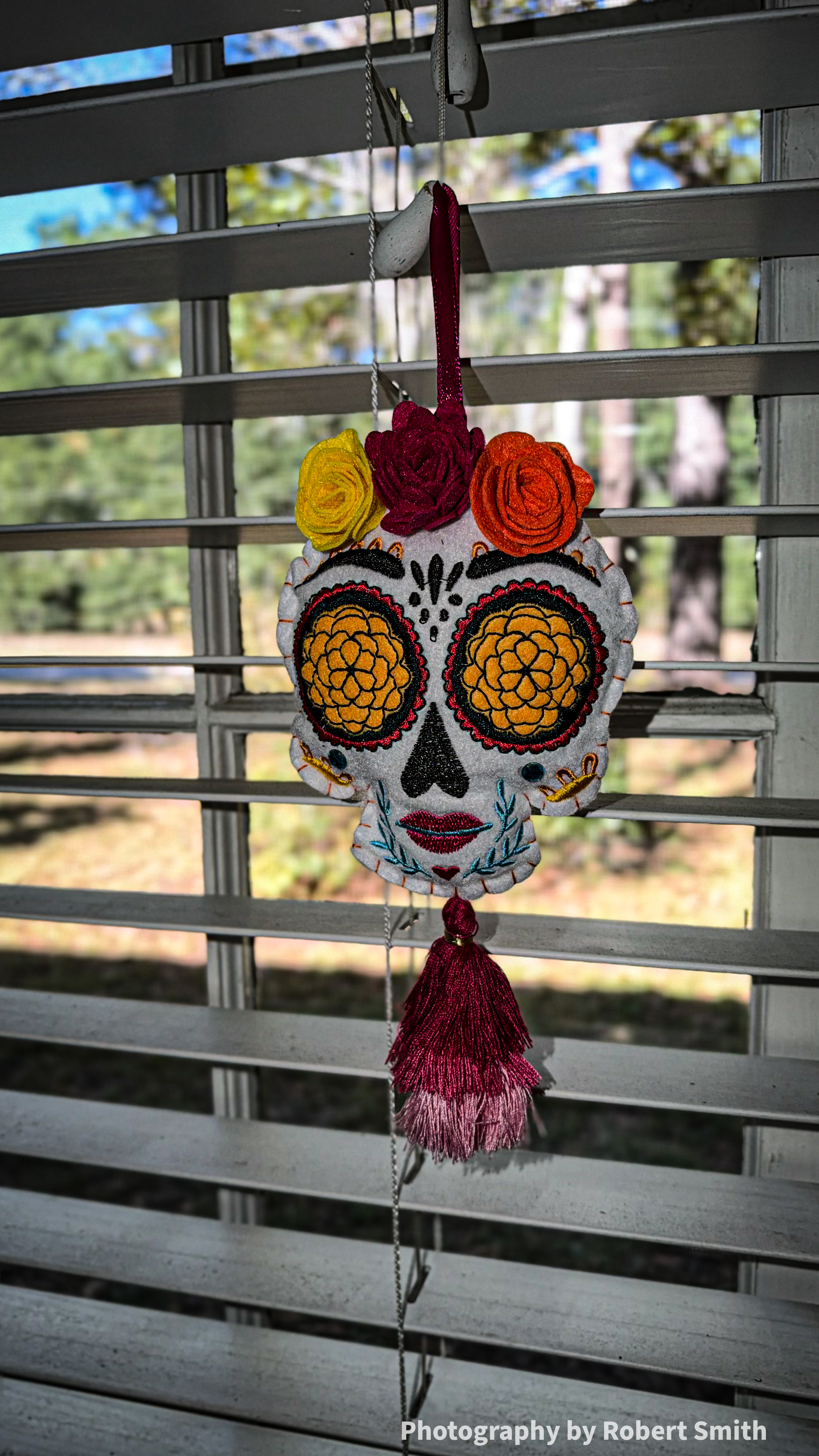 White embroidered skull ornament hanging from a burgundy ribbon in front of window blinds, with three fabric flowers above its face and a two-tone tassel below.