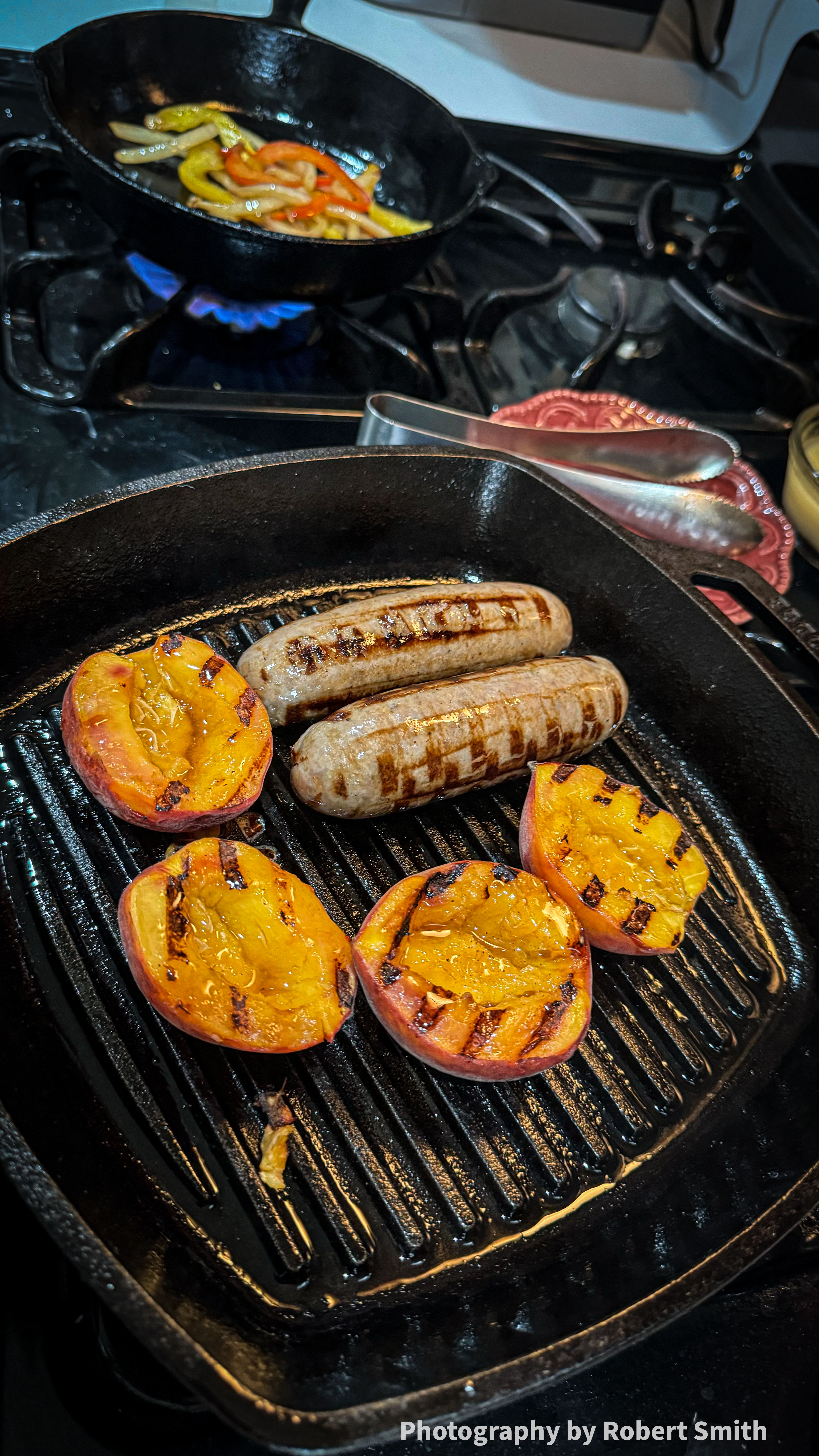 Bratwurst and peach halves in a ridged cast iron pan, with sliced peppers and onions cooking in a round skillet over a blue gas flame behind it.