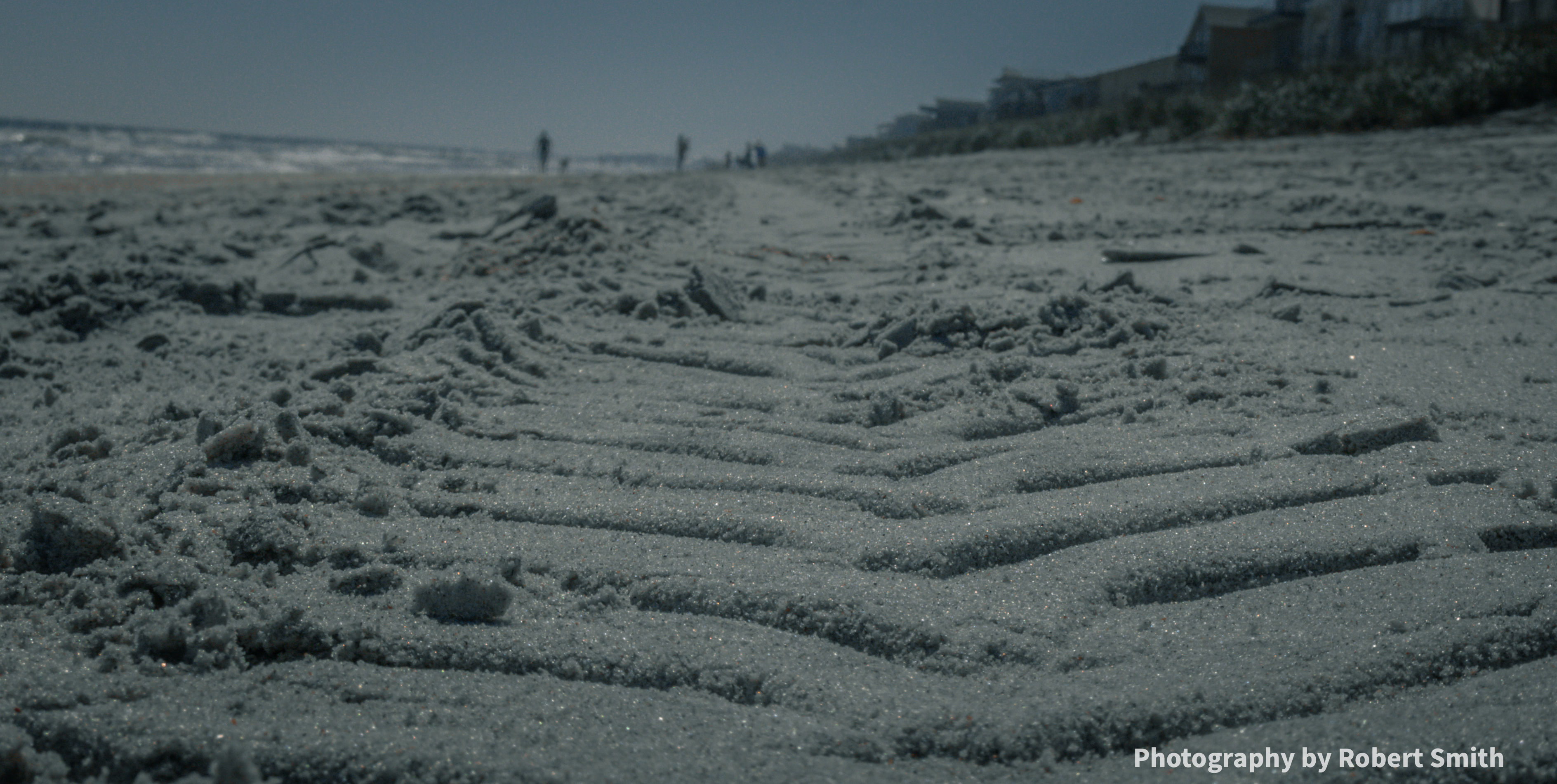 Close view of angled tire tread impressions and sparkling grains in beach sand, with the ocean, distant people, and beachfront houses blurred in the background.