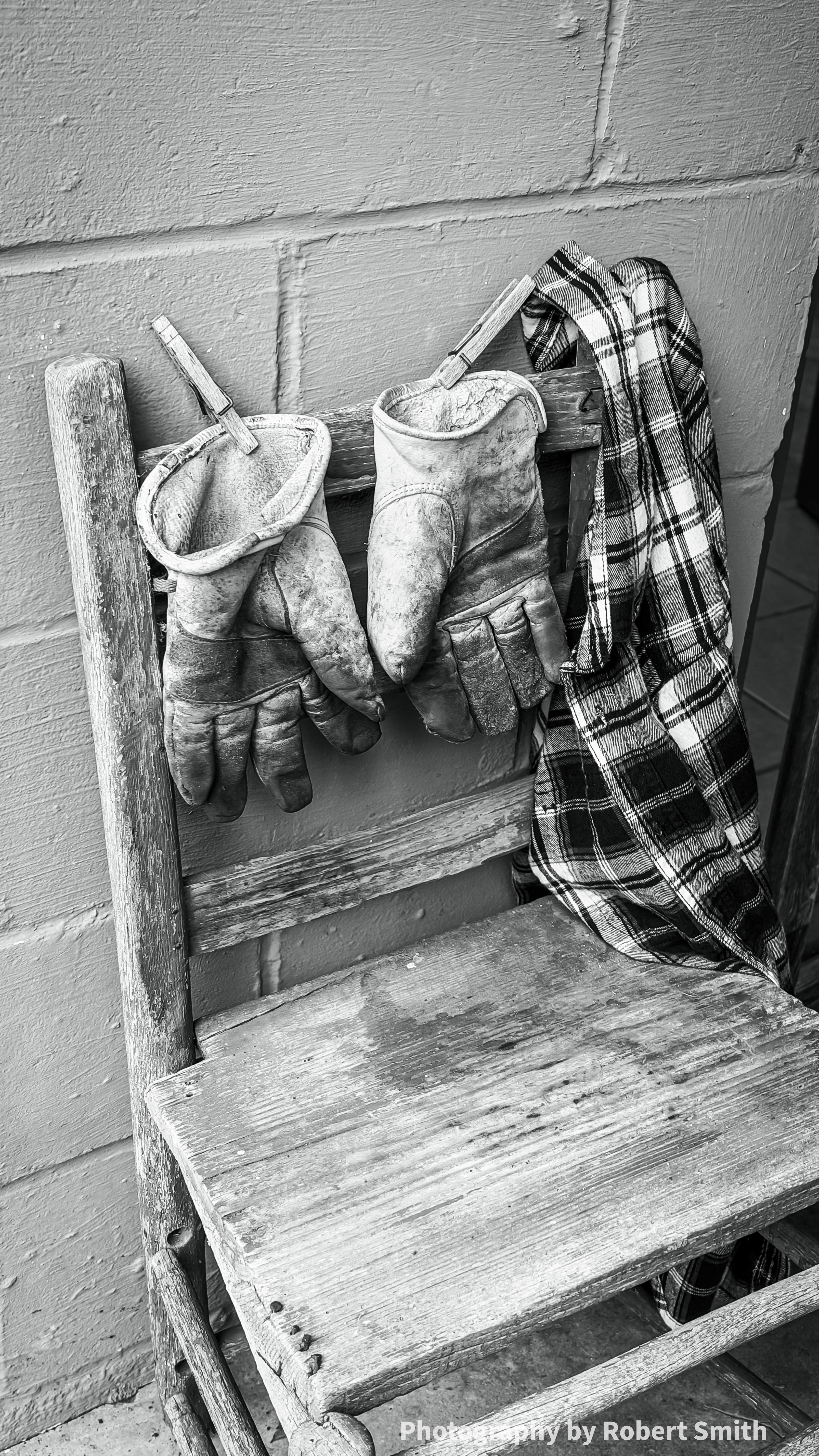 Angled black-and-white close view of two worn work gloves hanging from wooden clothespins on a chair back, beside a plaid shirt above the weathered seat.